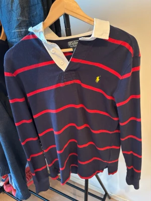 Randig tröja från Polo Ralph Lauren - Mörkblå långärmad tröja med röda ränder och vit krage från Polo Ralph Lauren. Klassisk gul logga broderad på bröstet och knappslå vid halsen. Perfekt för en sportig och avslappnad stil.