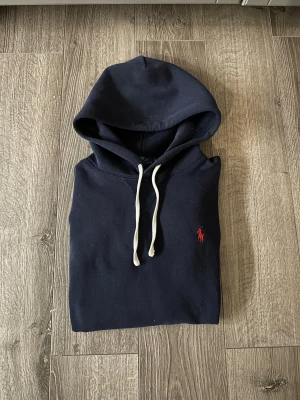 Mörkblå hoodie från Polo Ralph Lauren - Snygg mörkblå hoodie från Polo Ralph Lauren med klassisk huva och vita snören. Materialet är mjukt och bekvämt, perfekt för chill dagar. Ikonisk röd logga broderad på bröstet. Enkel och stilren design som är lätt att matcha med jeans eller joggers.