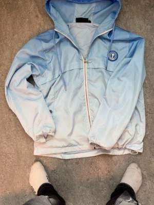 Ljusblå vindjacka från Trapstar - Ljusblå vindjacka från Trapstar med huva och dragkedja framtill. Jackan har elastiska muddar vid ärmslut och nederkant, samt logotyp på bröstet och Trapstar-tryck på ryggen. Materialet är lätt och passar perfekt för att skydda mot vind.