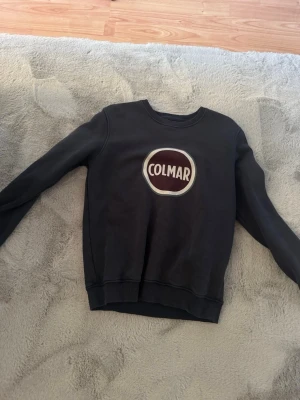 Svart Colmar sweatshirt med logga - Snygg svart sweatshirt från Colmar med stor vit och vinröd logga på bröstet. Tröjan har rund halsringning, långa ärmar och ribbade muddar. Perfekt för en avslappnad och trendig stil.