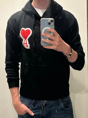 Svart hoodie från Ami Paris med hjärtdetalj - Säljer en svart hoodie från Ami Paris med stor röd hjärtdetalj och bokstaven A på bröstet. Hoodien har huva med dragsko, magficka och ribbade muddar. Perfekt för dig som gillar streetwear och vill ha något stilrent men ändå unikt.