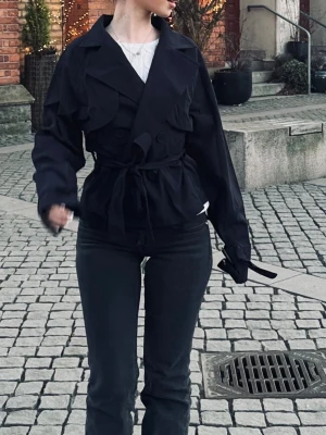 Svart trenchcoat med knytskärp - Stilren svart trenchcoat med dubbelknäppning och brett slag. Jackan har ett knytskärp i midjan och dekorativa detaljer på axlarna. Klassisk kortare modell som ger en cool och modern look. Perfekt för lager-på-lager och streetstyle.