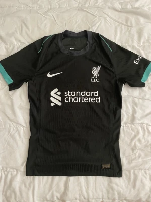 Svart Liverpool Nike matchtröja - Snygg svart Liverpool FC matchtröja från Nike med vita loggor och sponsortryck. Tröjan har rund hals, korta ärmar och turkosa detaljer på axlarna. Tillverkad i ett lätt och ventilerande funktionsmaterial som passar perfekt till träning eller match. Pris kan diskuteras 