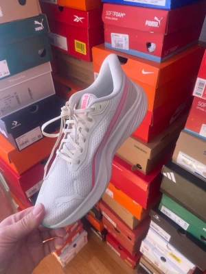Vita Puma sneakers med rosa detaljer - Snygga vita sneakers från Puma med meshmaterial och rosa detaljer längs sidan. Skorna har en sportig design med klassisk snörning och en tjock, bekväm sula. Perfekta för dig som gillar en fräsch och stilren look.