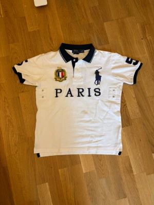 Vit pikétröja från Polo Ralph Lauren - Vit pikétröja från Polo Ralph Lauren med mörkblå detaljer, stor logga på bröstet, PARIS-tryck och broderad patch. Nummer 5 på ryggen och ärmarna. Klassisk krage och korta ärmar. Perfekt för en sportig och stilren look.