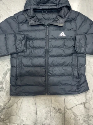 Svart dunjacka med huva från Adidas - Snygg svart dunjacka från Adidas med huva och dragkedja framtill. Jackan har Adidas-logga på bröstet och två sidofickor. Perfekt för kalla dagar och enkel att matcha med olika outfits.