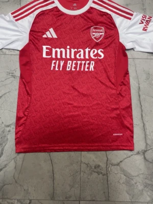 Arsenal matchtröja röd/vit Adidas - Snygg Arsenal matchtröja från Adidas i rött och vitt med klubbmärke och sponsortryck på bröstet. Tröjan har vita ärmar, röda axelränder och diskret mönster i tyget. Tillverkad i lätt och ventilerande material med AEROREADY-teknologi.