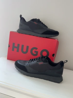 Svarta sneakers från Hugo - Snygga svarta sneakers från Hugo med diskreta röda detaljer och logga på sidan. Skorna har en modern siluett, snörning och är tillverkade i en mix av mocka och syntetmaterial. Perfekta för dig som vill ha en stilren och trendig look. Använd 1-2 gånger nypris 3,200 kr 