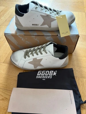 Golden goose sneaker - Snygga vita sneakers från Golden Goose med den ikoniska grå stjärnan på sidan och gråa skosnören. Skorna har svart detalj på hälen och en slitstark sula. Tillverkade i läder och mocka för en exklusiv känsla. Perfekta för dig som gillar streetstyle och vill sticka ut.