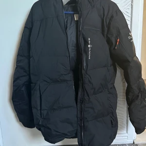 Svart dunjacka från Sail Racing - Säljer en svart dunjacka från Sail Racing med dragkedja framtill och ficka med orange dragkedja på ärmen. Jackan har en klassisk puffer-modell och är perfekt för kalla vinterdagar. Märkeslogga på bröstet och innerfoder i matchande färg. Hör av er för fler bilder.