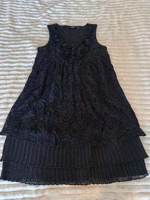 Svart spetsklänning från Isolde - Svart ärmlös klänning från Isolde med vacker spets, dekorativa blommor och pärlor vid ringningen. Klänningen har v-ringad hals och dubbla lager med plisserad nederdel som ger en elegant look. Perfekt för dig som gillar detaljer och stil.