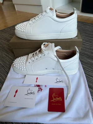 Vita Christian Louboutins - Snygga vita sneakers från Christian Louboutin med ikoniska röda sulor och coola vita nitar framtill. Skorna är i läder och har en klassisk låg siluett med vita snören. Perfekta för dig som vill sticka ut med exklusiv design och edgy detaljer.