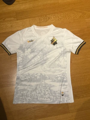 AIK Stockholm Edition tröja - Säljer en vit AIK matchtröja från Nike med snyggt stadsmotiv i ljusgrått på framsidan. Tröjan har AIK:s klubbmärke på bröstet och gulddetaljer på ärmsluten. Materialet är lätt och andas, perfekt för träning eller match.