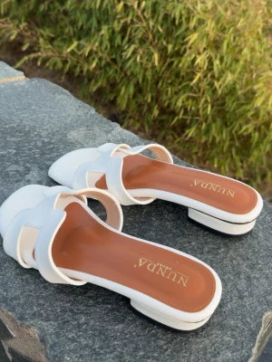 Vita sandaler från NUNDA - Säljer ett par stilrena vita sandaler från NUNDA med öppen tå och breda remmar i syntetmaterial. Sandalerna har en låg klack och brunt innersula med logotyp. Perfekt för dig som vill ha en enkel och snygg för sommaren. 