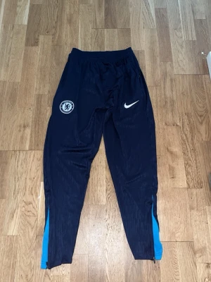 Chelsea träningsset från Nike - Säljer ett Chelsea träningsset från Nike, bestående av mörkblå långärmad tröja med ljusblå detaljer och matchande träningsbyxor. Tröjan har Bingx-logga och klubbemblem, byxorna har Nike-logga och Chelsea-emblem. Perfekt för fotbollsträning.