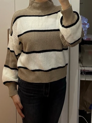 Randig stickad tröja med polokrage - Säljer en stickad tröja med polokrage i beige, vitt och svarta ränder. Tröjan har avslappnad passform, breda ärmar och ribbstickade muddar. Perfekt för dig som gillar en chill och trendig look.