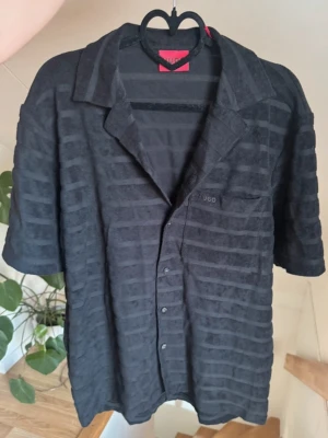 Svart skjorta från Hugo med struktur - Svart skjorta från Hugo med diskret randigt mönster i strukturen. Skjortan har korta ärmar, klassisk krage och knappar framtill. Tillverkad i ett mjukt material som ger en avslappnad känsla. Perfekt för dig som gillar stilrena plagg med en twist.