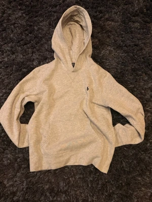 Grå hoodie från Ralph lauren - Snygg grå hoodie från Polo Ralph Lauren med klassisk huva och känguruficka . Tillverkad i mjukt sweatshirtmaterial som är riktigt skönt. Använd max 10 gånger och säljer pga av att den är för liten. Storlek L på barn storlekar och 14