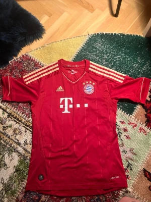 Bayern München Lahm matchtröja - Röd Bayern München matchtröja från Adidas med guldiga detaljer och klubbmärke på bröstet. Nummer 21 och Lahm på ryggen. Tröjan har korta ärmar, V-ringning och är tillverkad i lätt funktionsmaterial med CLIMACOOL-teknologi.