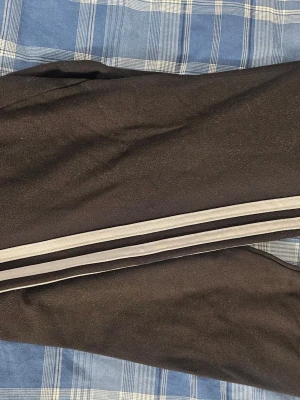 Svarta mjukisbyxor med vita ränder - Svarta mjukisbyxor med två vita ränder längs sidorna. Byxorna har en klassisk sportig look och är tillverkade i ett mjukt och bekvämt material, perfekt för chill dagar eller träning. Snygg och enkel design som passar till det mesta.