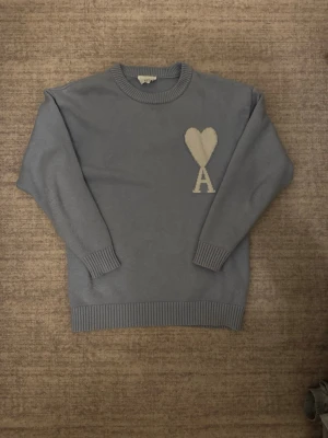 Ami Paris Tröja - Säljer en blå stickad tröja från Ami Paris med ett vitt hjärta och bokstaven A broderat på bröstet.  Tröjan har rund halsringning och ribbade muddar och nederkant.  En perfekt tröja för dig som uppskattar stilrena detaljer och klassisk design. 