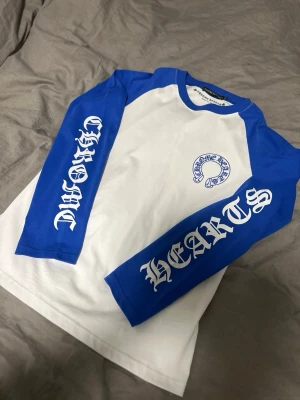 Blåvit långärmad topp från Chrome Hearts - Snygg långärmad topp från Chrome Hearts med blå ärmar och vit kropp. Stora tryck på ärmarna och ryggen i blått och vitt ger en cool streetwear-look. Perfekt för dig som gillar statement-plagg och unika detaljer. Storlek XxL(passar L)