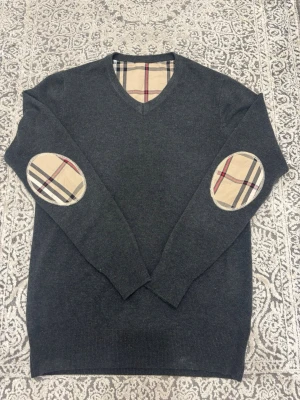 Mörkgrå stickad tröja Burberry - Mörkgrå stickad tröja från Burberry med V-ringning och klassiska rutiga detaljer vid armbågar och insidan av kragen. Tröjan har långa ärmar och ribbade muddar vid ärmslut och nederkant. Perfekt för dig som gillar stilrena och tidlösa plagg.