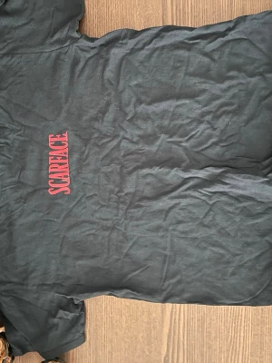 Svart Scarface t-shirt med tryck - Cool svart t-shirt med stort Scarface-tryck i rött och vitt både fram och bak. På ryggen finns en ikonisk bild från filmen och text. Perfekt för dig som gillar filmklassiker och vill sticka ut med en unik t-shirt.