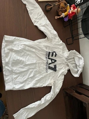 Vit hoodie från Emporio Armani EA7 - Snygg vit hoodie från Emporio Armani EA7 med stor logga på bröstet. Hoodien har huva och en klassisk känguruficka framtill. Perfekt för en avslappnad och trendig stil.
