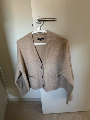 Beige kofta från Uniqlo - Mysig beige kofta från Uniqlo med v-ringad hals, tre knappar framtill och två fickor. Koftan har ribbade muddar och är tillverkad i ett mjukt stickat material som passar perfekt för lager på lager.