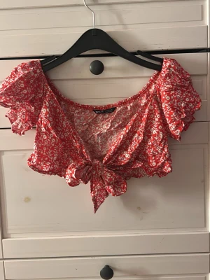 Röd blommig croppad blus med knyt - Supersöt röd blus med vita småblommor, croppad passform och knyt framtill. Toppen har korta puffärmar och en luftig, somrig känsla. Perfekt till höga jeans eller kjol.❤️