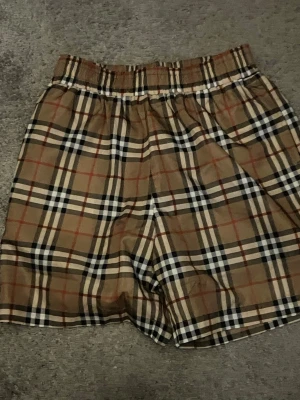 Rutig Burberry shorts i bomull - Säljer ett par klassiska rutig shorts från Burberry i beige, svart, vit och röd. De har elastisk midja och är tillverkade i 100% bomull, vilket gör dem riktigt sköna och lätta att bära. Perfekt för dig som gillar ikoniska mönster och stilrena plagg.Burberry swim shorts storlek S