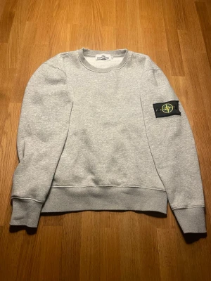 Grå sweatshirt från Stone Island - Säljer en stilren grå sweatshirt från Stone Island med klassisk rund hals och ribbade muddar. Tröjan har den ikoniska svarta patchen med logga på vänster ärm. Tillverkad i mjuk bomullsblandning och har en clean look som funkar till allt.