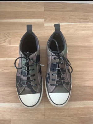 Valentino camo sneakers med VLTN-snören Storlek 43 - Snygga sneakers från Valentino i canvas med camouflagemönster i grönt, beige och svart. Skorna har vita tåpartier, svarta VLTN-loggade skosnören och svarta detaljer. Perfekta för dig som vill sticka ut med en exklusiv streetstyle-look. Storlek 43, mycket bra kvalite men har används. Skobox ingår ej men däremot ett extra skösnöre. Pris kan diskuteras,hör av dig!