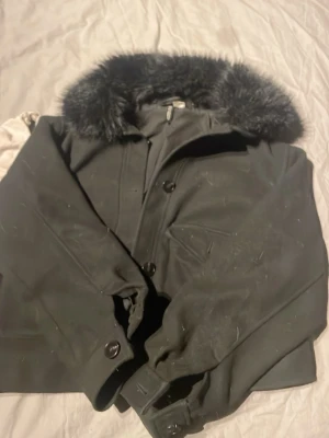 Meotine liknande jacka - Stilren svart kappa från H&M Divided med stor krage i svart fuskpäls. Kappan har knäppning framtill, fickor och snygga detaljer vid ärmsluten. Perfekt för kalla dagar när du vill vara både varm och trendig.