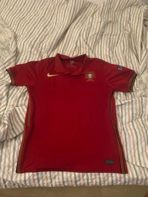 Portugal Nike fotbollströja röd - Röd Portugal fotbollströja från Nike med broderad landslagssköld på bröstet och gul Nike-logga. Tröjan har krage, korta ärmar med randiga detaljer och märken på båda ärmarna. Perfekt för fotbollsfans och matcher.