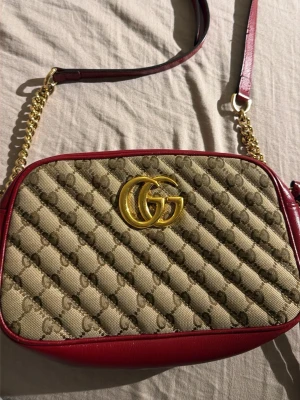 Gucci GG Marmont axelväska röd/beige - Snygg Gucci GG Marmont axelväska i beige canvas med GG-mönster och röda läderdetaljer. Väskan har guldfärgad kedja och GG-logo framtill. Insidan är fodrad i beige mocka och har dragkedja upptill. Perfekt för dig som vill ha en trendig och lyxig väska.