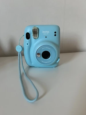 Blå Fujifilm Instax Mini 11 kamera - Säljer en ljusblå Fujifilm Instax Mini 11 direktfilmskamera. Kameran har en rundad form, inbyggd blixt och selfie-spegel på framsidan. Tillverkad i plast och har en matchande handledsrem. Perfekt för att ta instant-bilder med vänner.