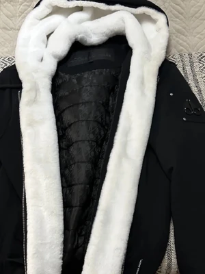 Moose knuckles kofta/jacka - Snygg svart kappa från Moose Knuckles med stor huva fodrad i vit fuskpäls. Insidan har quiltat mönster och jackan har en klassisk, stilren look. Perfekt för kalla dagar när du vill vara både varm och trendig.