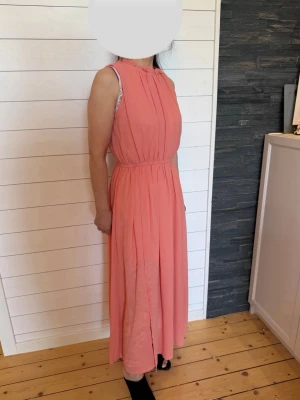 Lång korallrosa ärmlös klänning - Helt ny: Snygg långklänning i korallrosa med ärmlös design och lätt rynkad halsringning. Klänningen har markerad midja och ett luftigt, fallande tyg som ger en elegant siluett. Perfekt för sommarens tillfällen.