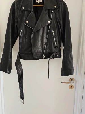 Svart skinnjacka från Tango - Snygg svart skinnjacka från Tango med klassisk bikerstil, sned dragkedja, silvriga detaljer och bälte i midjan. Jackan har flera fickor med dragkedja och coola detaljer som ger en edgy look.