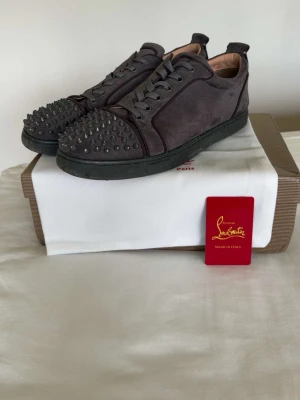 Gråa Louboutin sneakers med nitar - Snygga gråa sneakers från Christian Louboutin i mocka med ikoniska nitar på tån och svarta snören. Skorna har den klassiska röda sulan och en låg profil. Perfekta för dig som vill sticka ut med en edgy och lyxig stil. Storlek 46, mycket bra skick! 