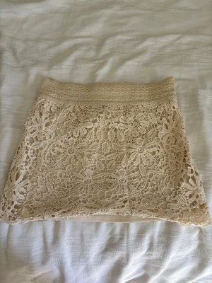 Beige spetskjol med virkad midja - Söt beige kortkjol med vacker spets och virkad midja. Kjolen har ett blommigt spetsmönster och är fodrad undertill. Perfekt för dig som gillar romantisk och feminin stil. Kjolen är aldrig använd och är i superbra skick! Tveka inte att höra av dig ifall du har frågor!🫶🏻