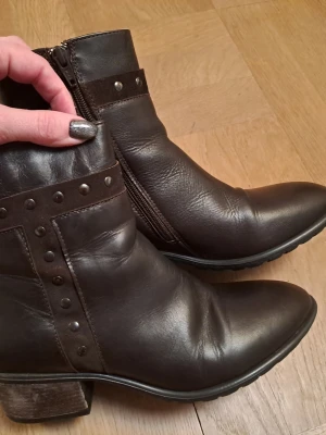 Bruna boots med nitar och dragkedja - Snygga bruna stövletter i skinn med dekorativa nitar och dragkedja på sidan. Boots med låg klack och rundad tå, perfekta för en cool och trendig look. Materialet är mjukt läder och detaljerna i mocka ger extra stil.