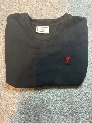 Svart t-shirt från Ami med röd broderi - Svart t-shirt från Ami med ett litet rött broderat hjärta och bokstaven A på bröstet. Klassisk rund halsringning och korta ärmar. Tillverkad i mjuk bomull som känns skön mot huden. Perfekt för en clean och stilren look.