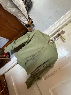 Grön hoodie med volangdetaljer - Säljer en ljusgrön hoodie med dragkedja och volangdetaljer längs ärmarna. Tröjan har ribbade muddar och en enkel, stilren design utan tryck. Perfekt för dig som gillar en unik twist på klassiska hoodies. För barn 10-11år