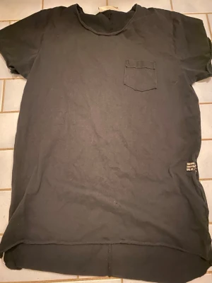 Svart t-shirt med bröstficka - En svart t-shirt med rund hals och en liten bröstficka på vänster sida. T-shirten har korta ärmar och en något längre bakdel. Perfekt för en avslappnad och enkel stil.