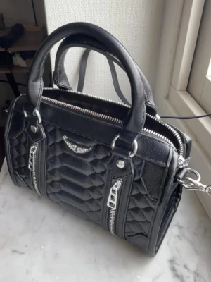 Svart väska med silver detaljer - Söt väska med silverdetaljer, säljer då den inte kommer till användning.                                           Den är i oanvänt skick!                                            Den har axelband som går att ta av.                                Köpt på marknad utomlands.                                              Nypris 500💗