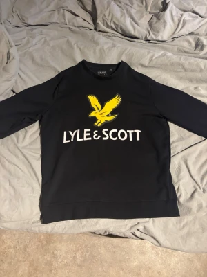 Svart sweatshirt från Lyle & Scott - Säljer en svart sweatshirt från Lyle & Scott med stort gult örntryck och vit logga på bröstet. Tröjan har rund halsringning och långa ärmar. Perfekt för dig som gillar streetwear och tydliga loggor.
