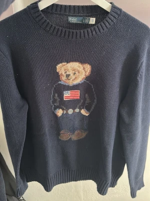 Ralph Lauren stickad tröja - Mörkblå stickad tröja från Polo Ralph Lauren med ikoniska Polo Bear-motivet framtill. Säljer tröjan för jag inte använder den längre. Storlek L men sitter som M. Säljs för 750kr Kontakta mig om du har frågor.